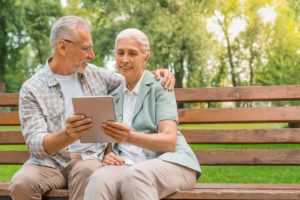 Happy retired couple sitting at park bench and using digital tablet